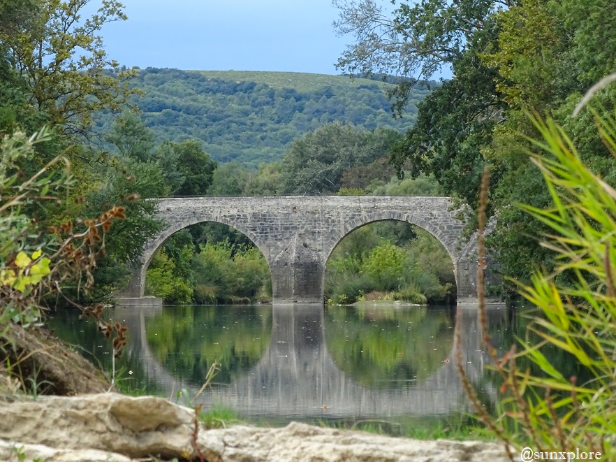Un pont en pierre à deux arches se reflète dans l'eau de la Cèze, entouré par la nature.